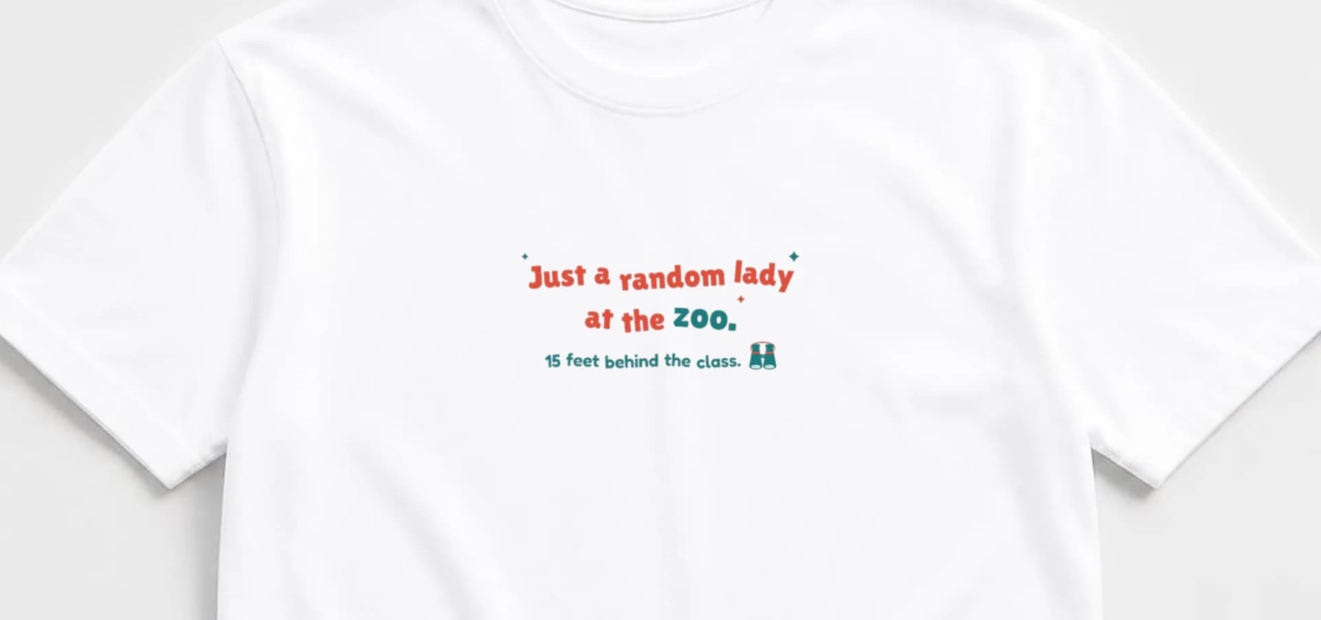 Close-up of white tee with "Just a random lady at the zoo." in coral and teal bouncy lettering and a tiny pair of binoculars