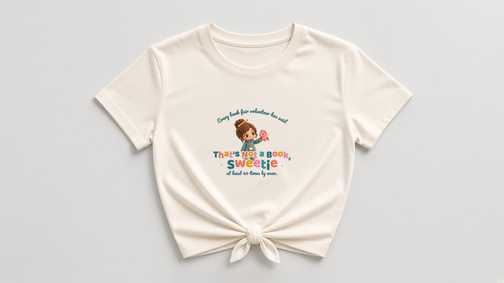 Natural cream knotted women's shirt with a plush chibi volunteer holding up a pink unicorn plush, under "Every book fair volunteer has said" in teal script, with multicolor "That's Not a Book, Sweetie" lettering, "at least 40 times by noon." and tiny gold sparkles