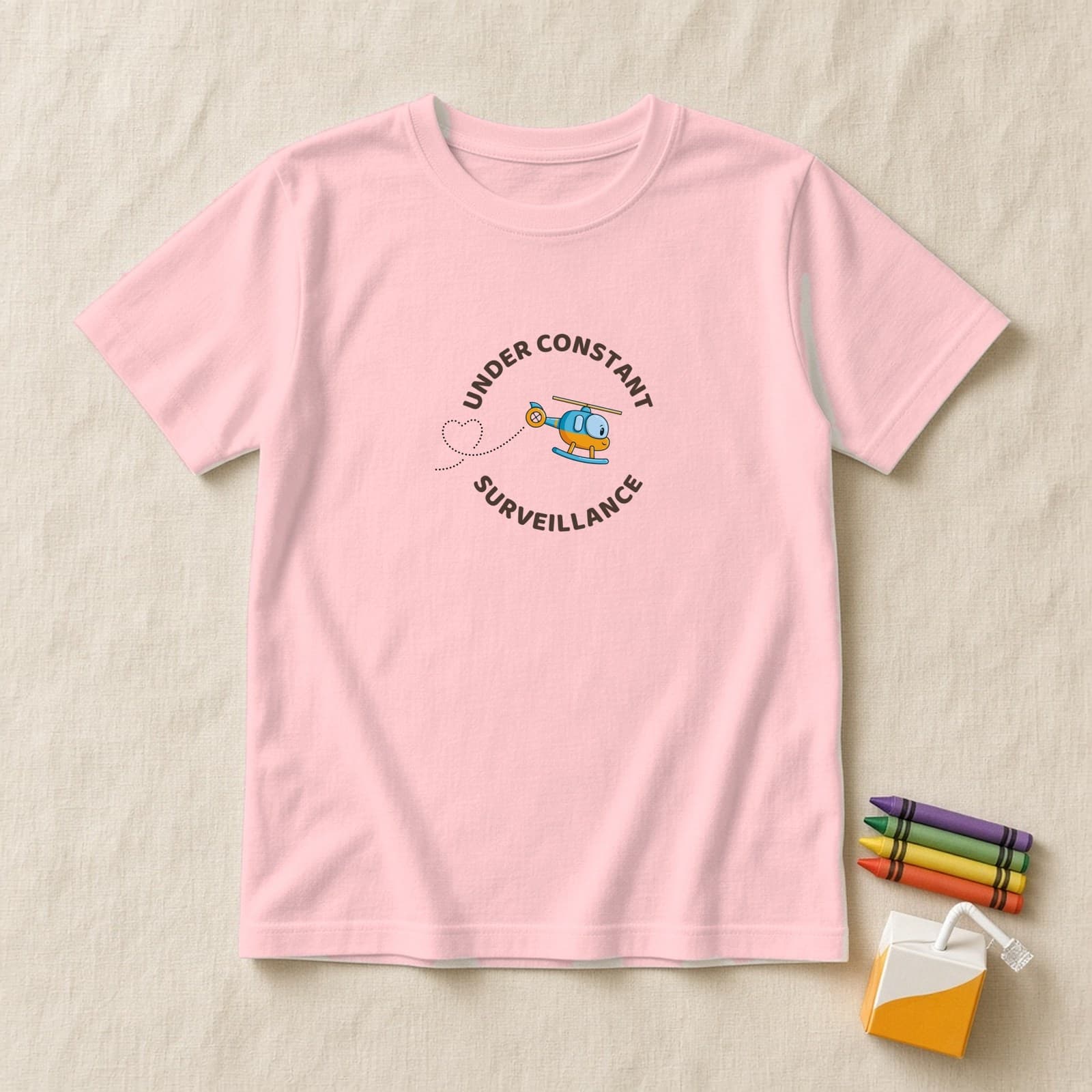 Pink kids shirt with a round Under Constant Surveillance badge and a tilted cute helicopter with a heart trail, flat lay on cream linen with crayons