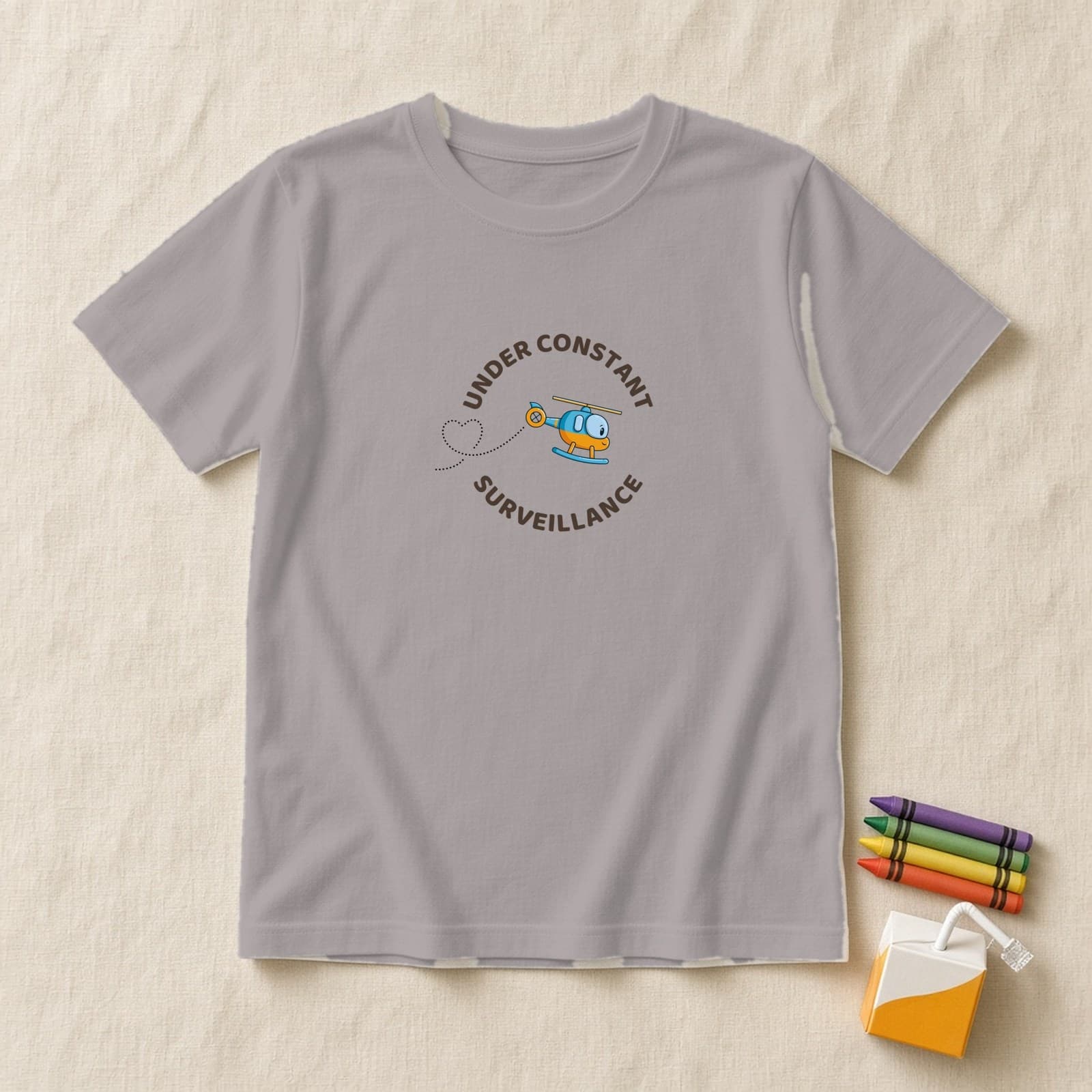 Athletic Heather kids shirt with a round Under Constant Surveillance badge and a tilted cute helicopter with a heart trail, flat lay on cream linen with crayons