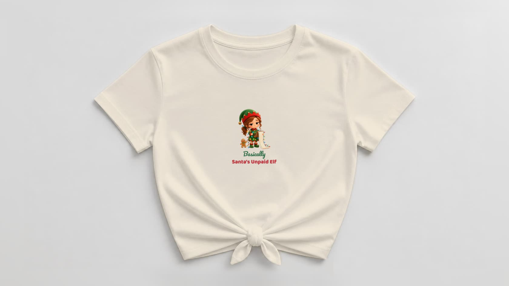 Natural cream knotted women's tee with a deadpan plush elf mom wrapped in Christmas lights and holding an endless checklist, a gingerbread sidekick at her feet, and "Basically Santa's Unpaid Elf" in green and red lettering