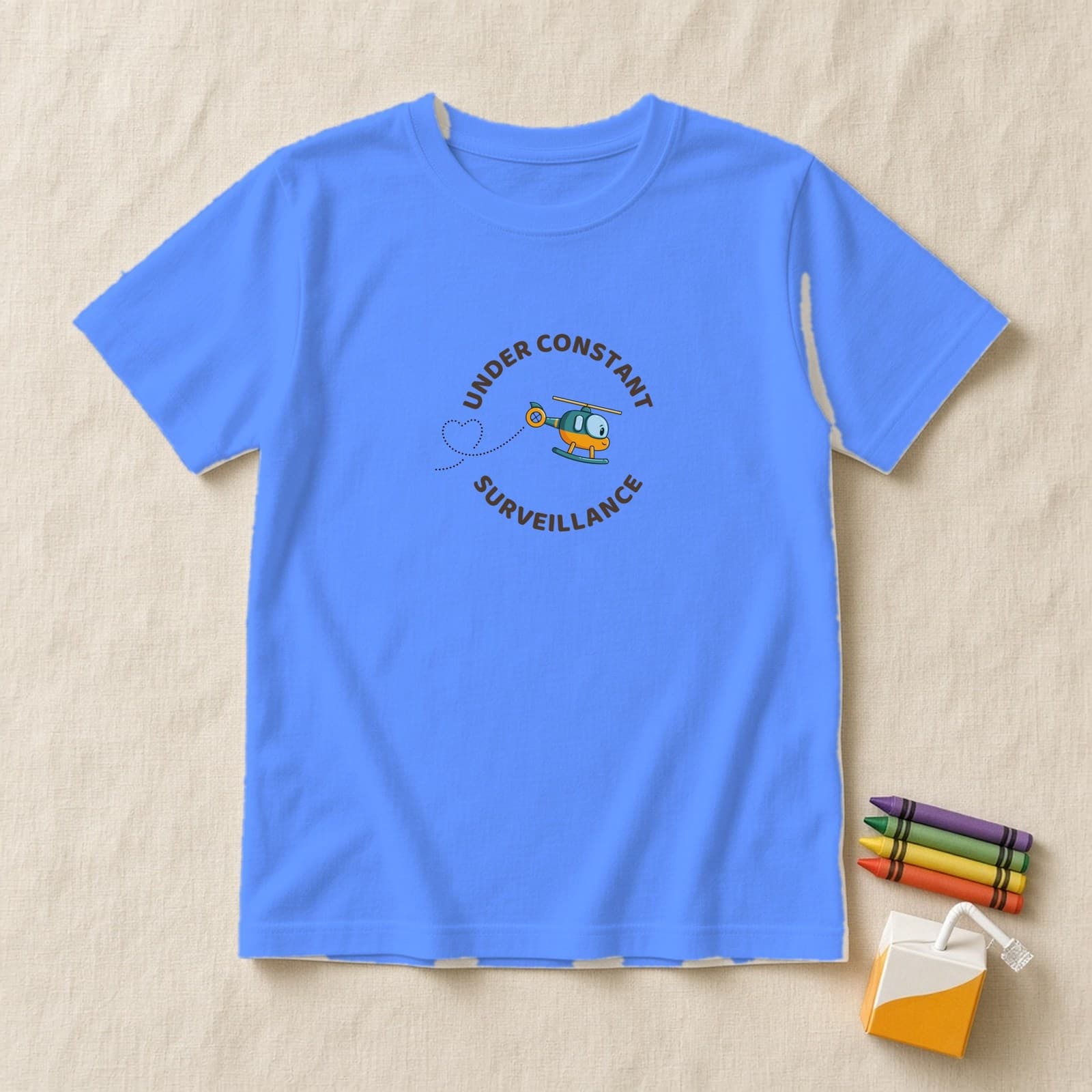 Heather Columbia Blue kids shirt with a round Under Constant Surveillance badge and a tilted cute teal helicopter with a heart trail, flat lay on cream linen with crayons