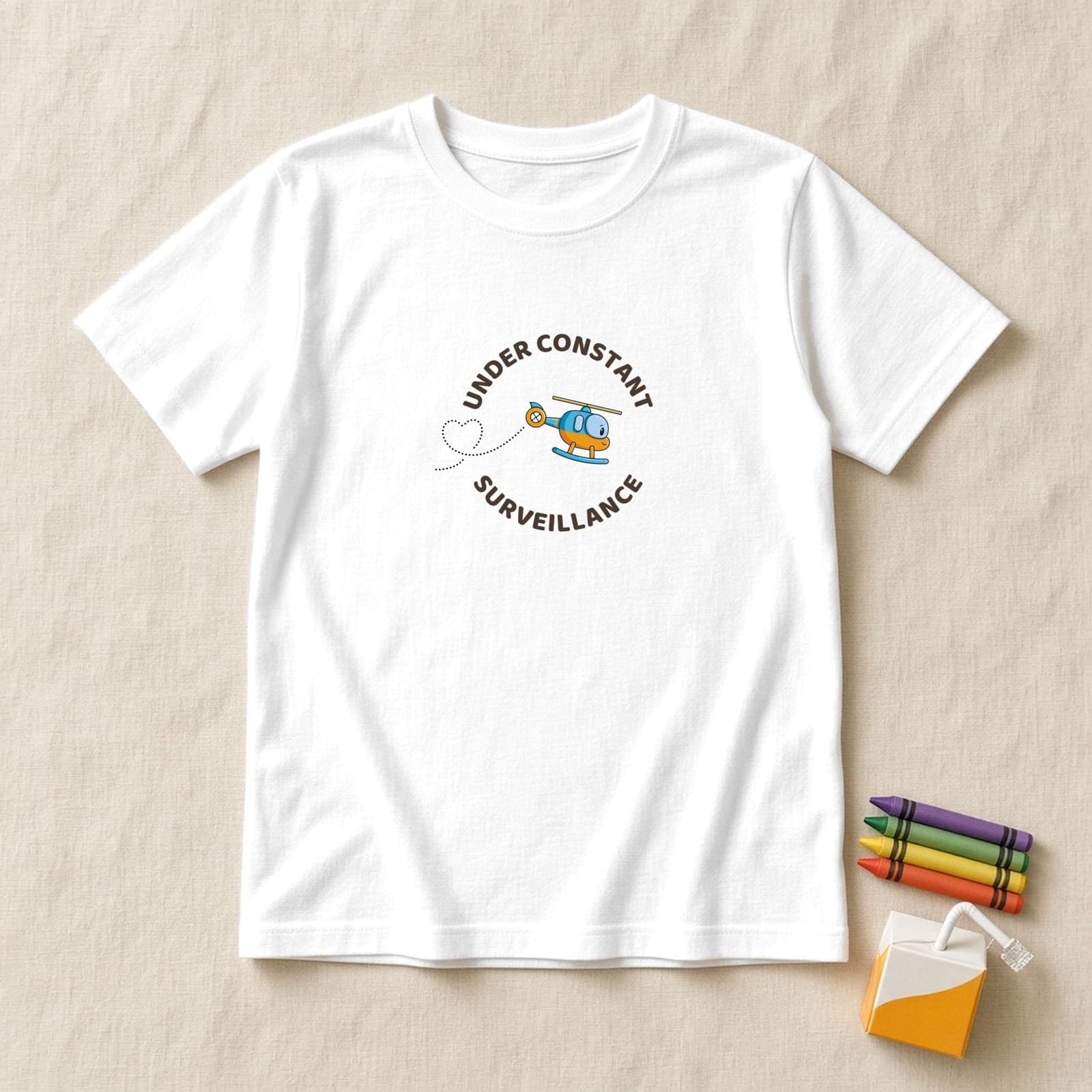 White kids shirt with a round Under Constant Surveillance badge and a tilted cute helicopter with a heart trail, flat lay on cream linen with crayons