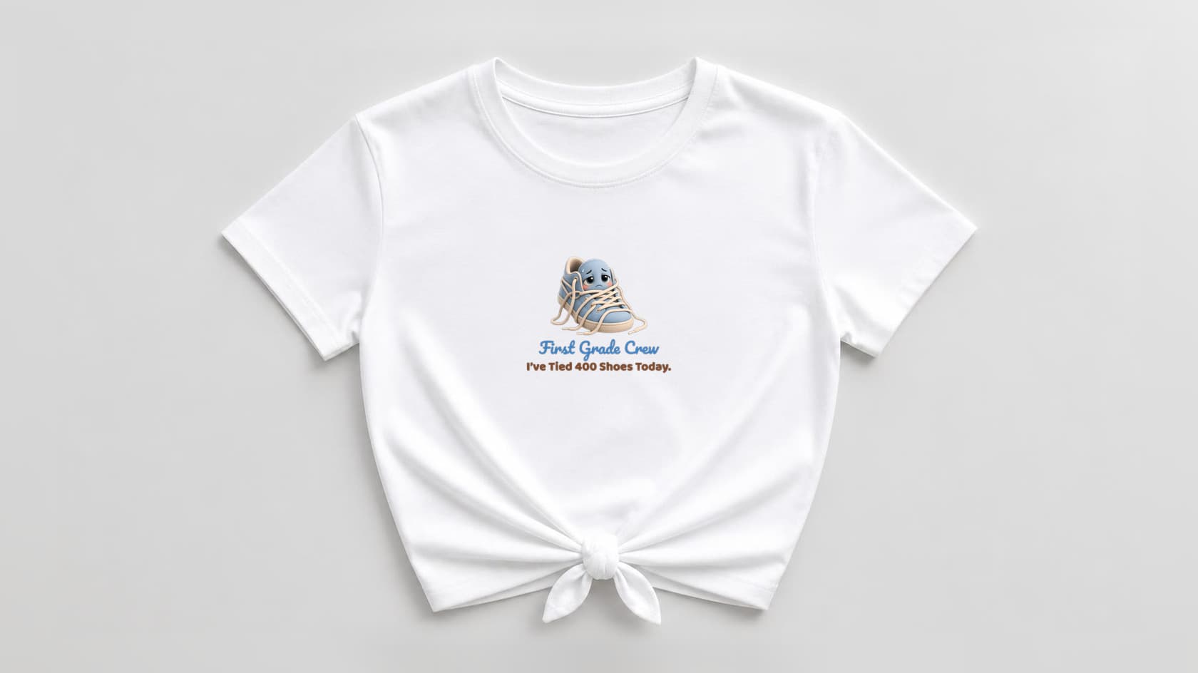 White knotted women's shirt with a worried plush blue sneaker tangled in long cream laces, above "First Grade Crew" in sky blue script and "I've Tied 400 Shoes Today." in bold brown