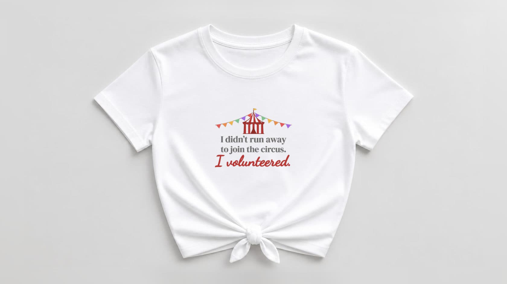White knotted tee with a red-and-white circus big top and pastel rainbow bunting, "I didn't run away to join the circus." in warm gray serif and "I volunteered." in red script