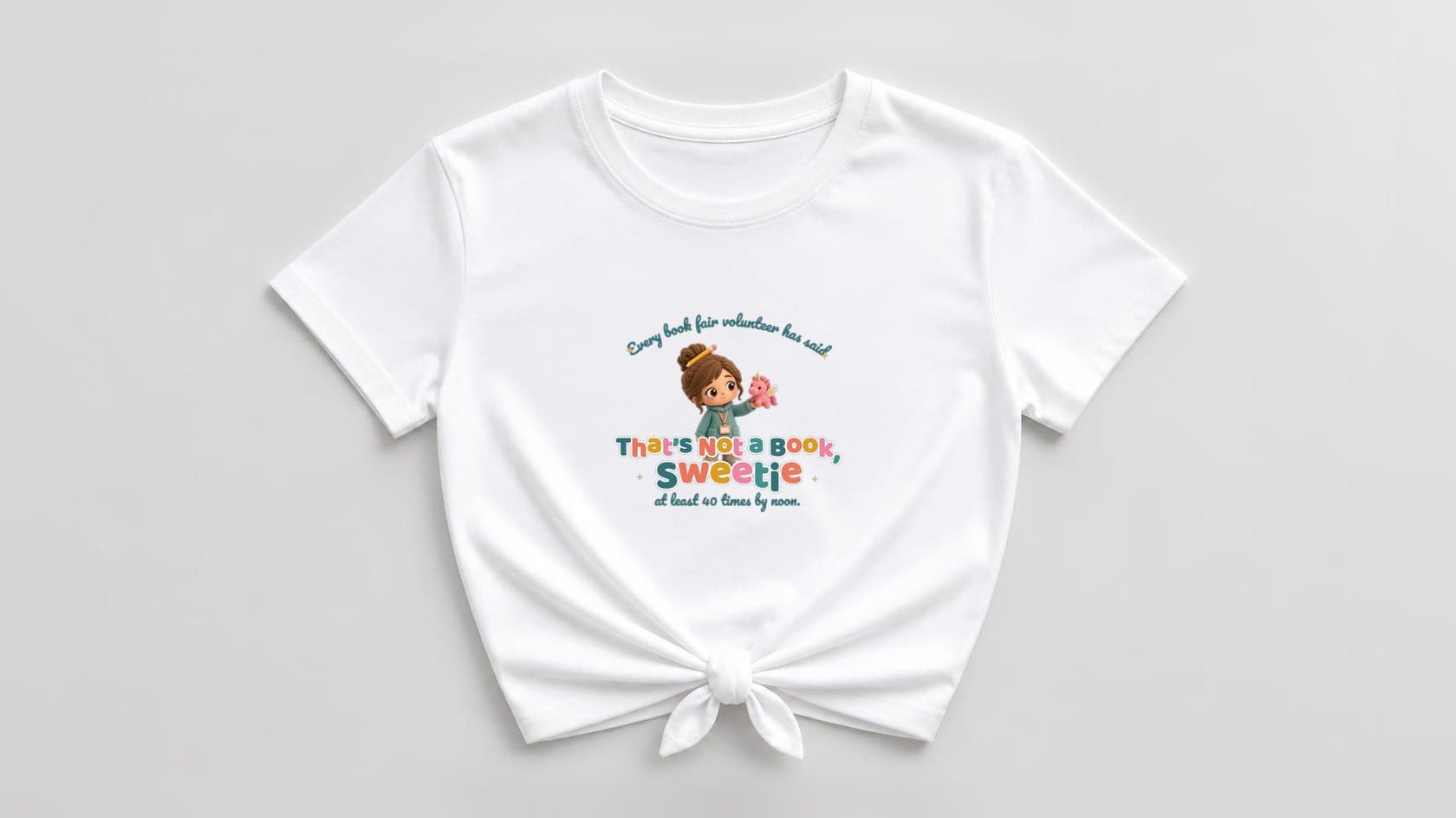 White knotted women's shirt with a plush chibi book fair volunteer in a teal sweater, pencil in her messy bun and a peach lanyard, holding up a pink unicorn plush, under "Every book fair volunteer has said" arched in teal script, with a big multicolor "That's Not a Book, Sweetie" in rounded letters and "at least 40 times by noon." below